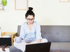Woman sitting on a couch working on a computer holding a coffee mug in her hand. 